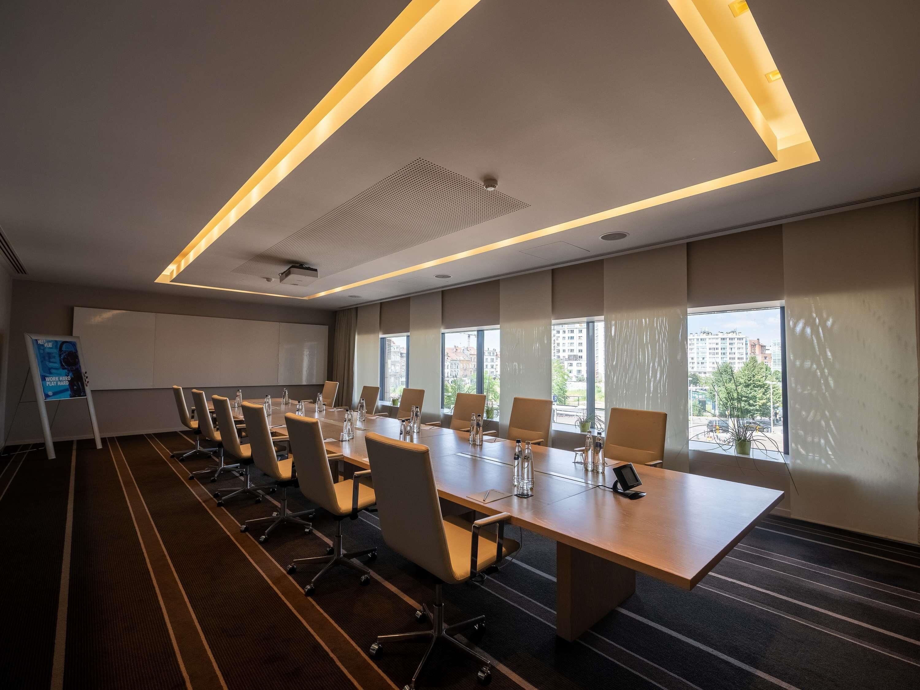 Ein Konferenzraum mit einem langen Tisch, Stühlen, einer Projektionsleinwand und großen Fenstern mit Blick auf die Stadt.