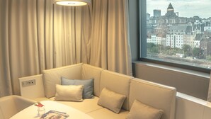 A modern room with a sofa, a small table, and a window with a city view. - Pullman Brussels Centre Midi (Brussels)