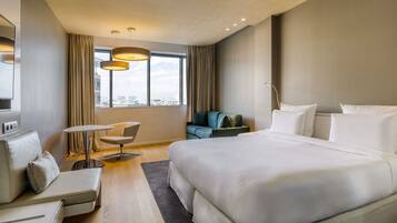 Une chambre d’hôtel moderne avec un grand lit, un canapé, une petite table et une chaise. Une grande fenêtre, ornée de rideaux, offre une vue sur la ville.