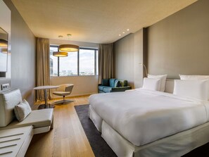Camera d'albergo moderna con un letto grande, un divano, un tavolino e una sedia. È presente un'ampia finestra con tende, e si può ammirare una vista sulla città.
