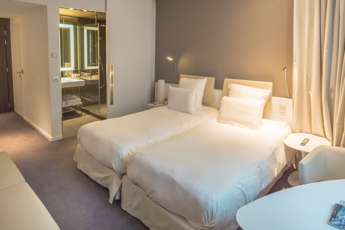 Habitación de hotel con una cama grande, una mesita de noche, una silla y un baño con cabina de ducha de vidrio.