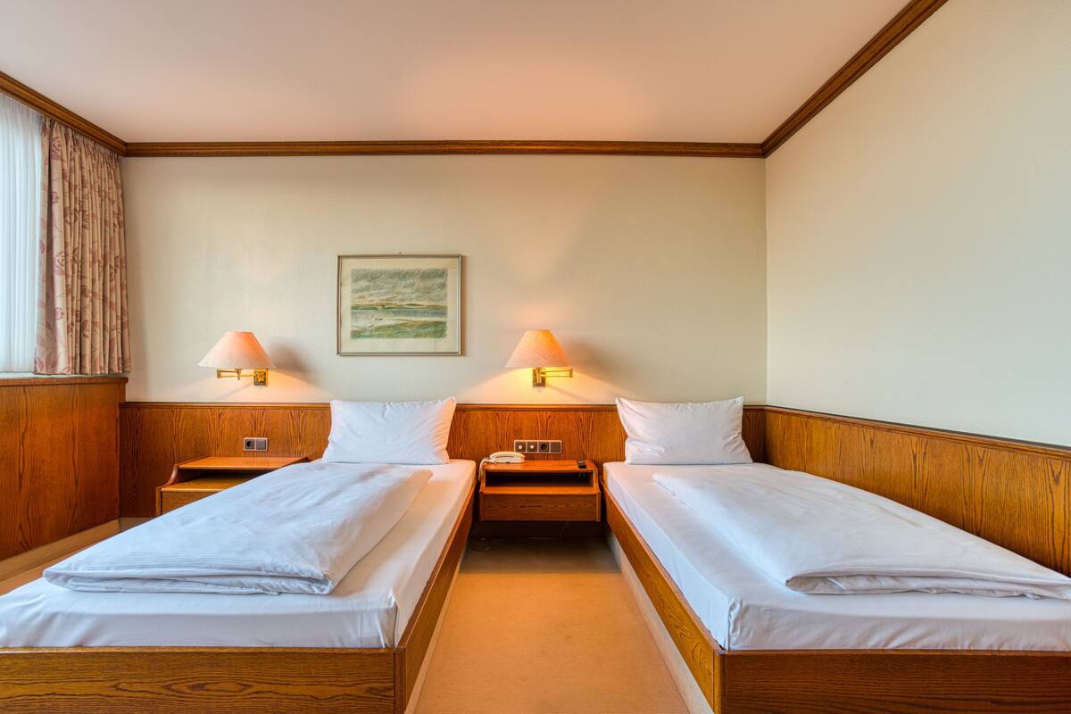 A hotel room with two single beds, wooden headboards, bedside tables with lamps, and a framed picture on the wall.