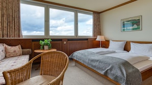 A hotel room with a large window, a bed, a sofa, a chair, and a small table with a plant. - FF&E Hotel Dania (Fehmarn)