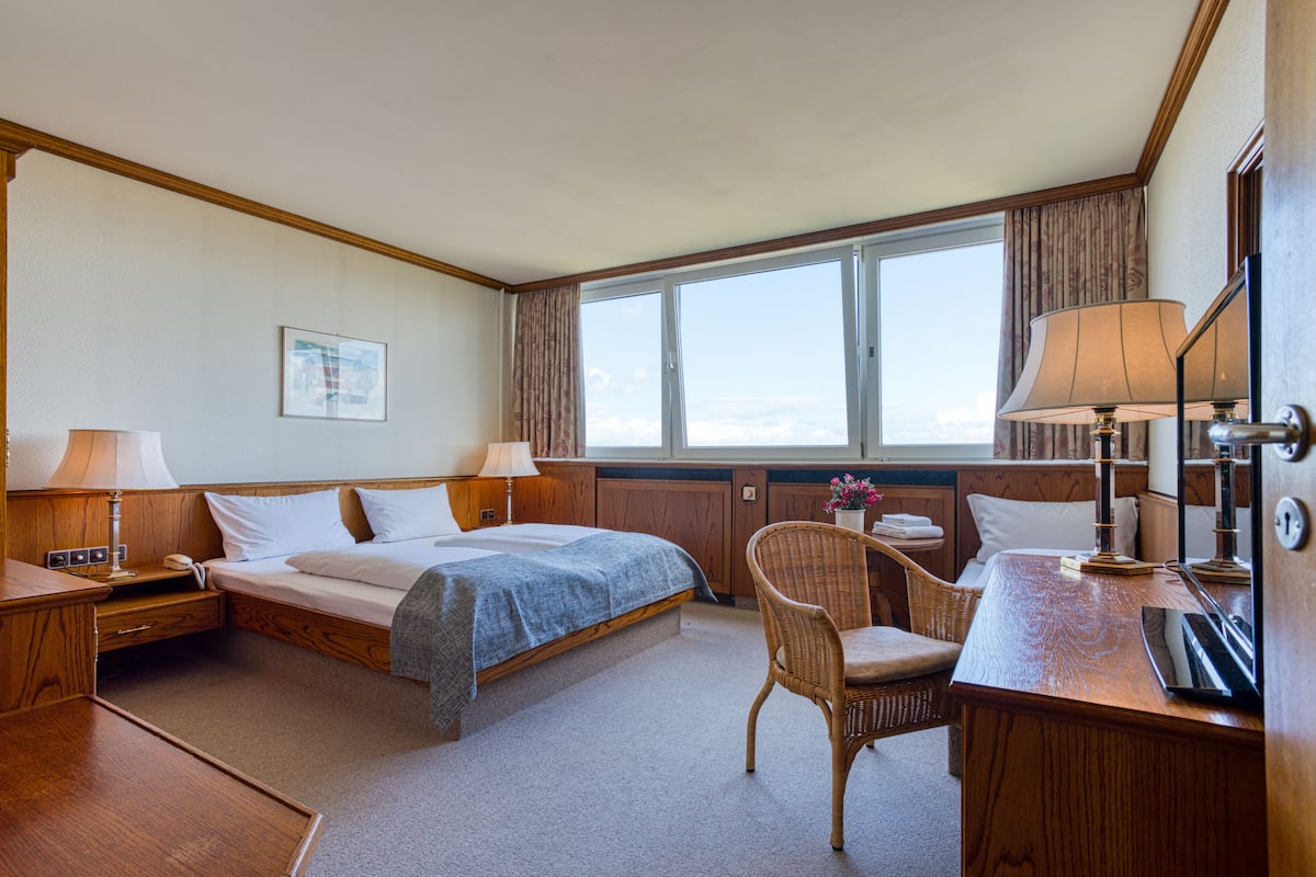 A hotel room with a bed, two bedside tables, a desk, a chair, and a large window with curtains.