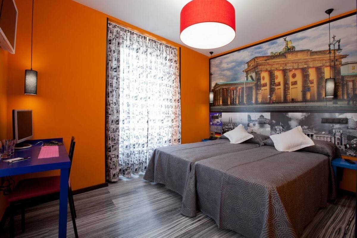 Habitación de hotel con dos camas, un escritorio, una silla, una lámpara colgante roja y un gran mural con vista a la ciudad.