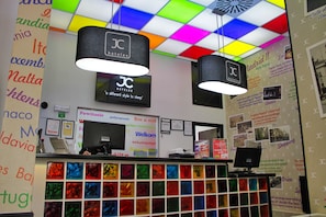 A hotel reception area with colorful ceiling tiles, black hanging lamps with 'JC Hotels' branding, a computer desk, and a wall with multilingual welcome signs and travel information. - Jc Rooms Santo Domingo (Madrid)