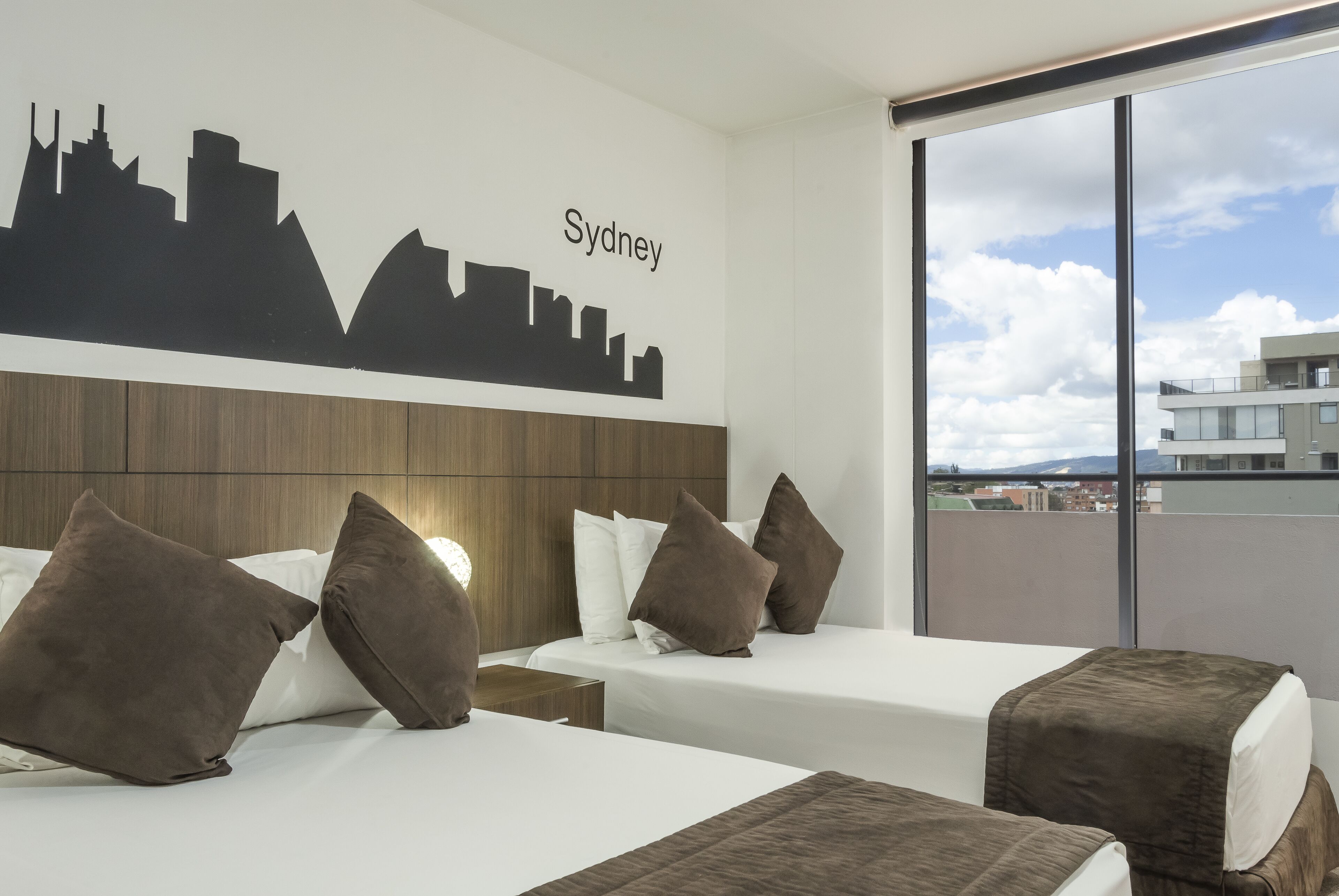 Une chambre d’hôtel avec deux lits, une fresque murale représentant le paysage urbain et une grande fenêtre donnant sur les bâtiments.