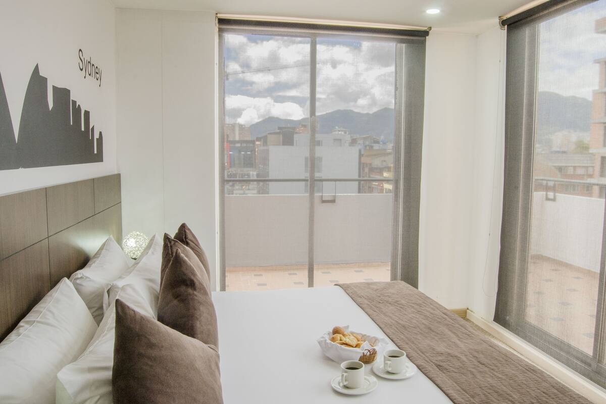 A hotel room with a bed, pillows, a table with a basket of pastries and cups, and a large window with a cityscape view.