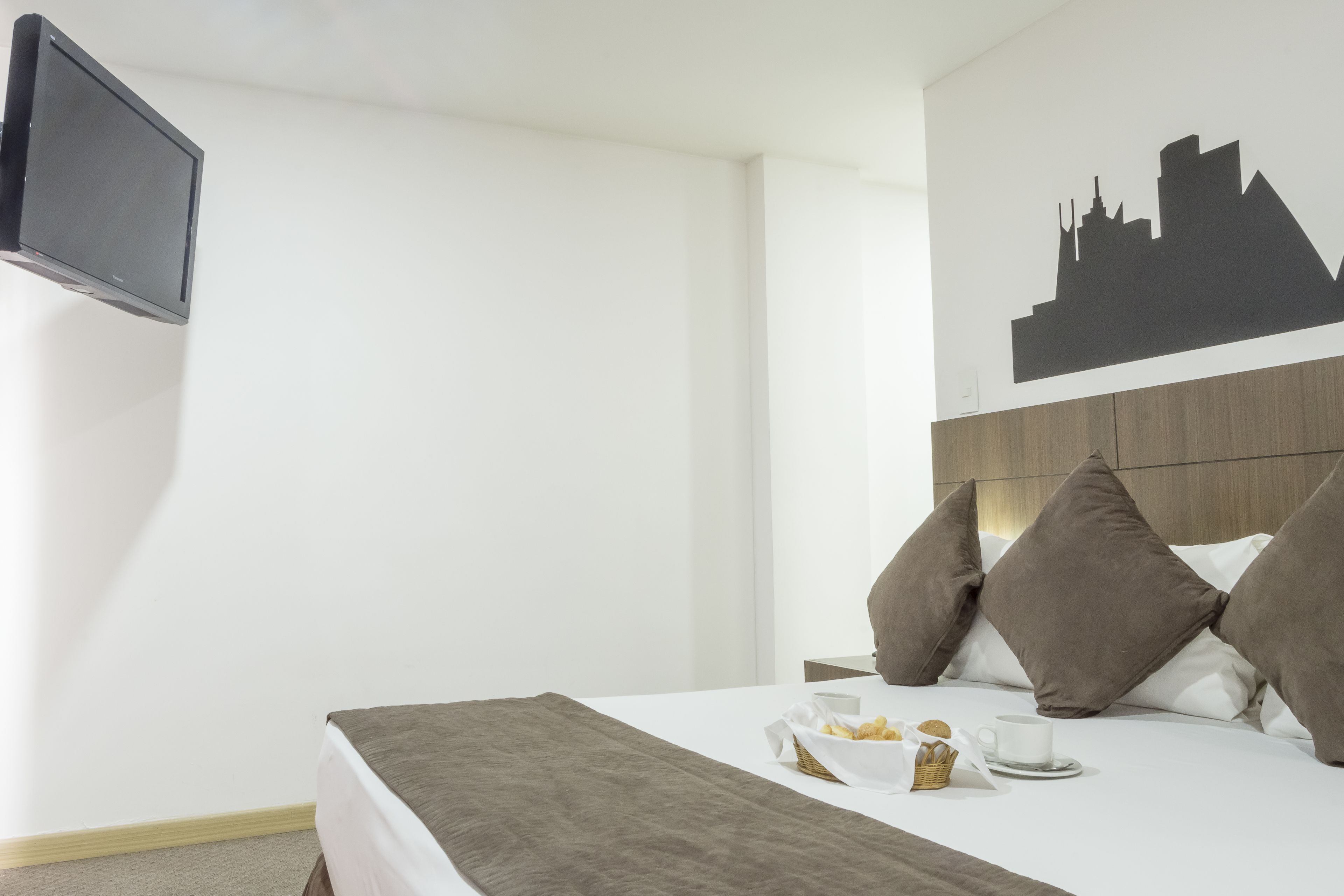 Une chambre d’hôtel avec un lit, un téléviseur à écran plat fixé au mur, une œuvre d’art représentant un paysage urbain au-dessus du lit et un plateau de petit-déjeuner sur le lit.
