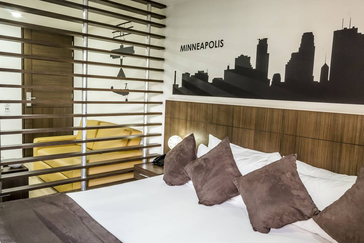 A hotel room with a bed, a city skyline mural, a wooden headboard, and a yellow chair.
