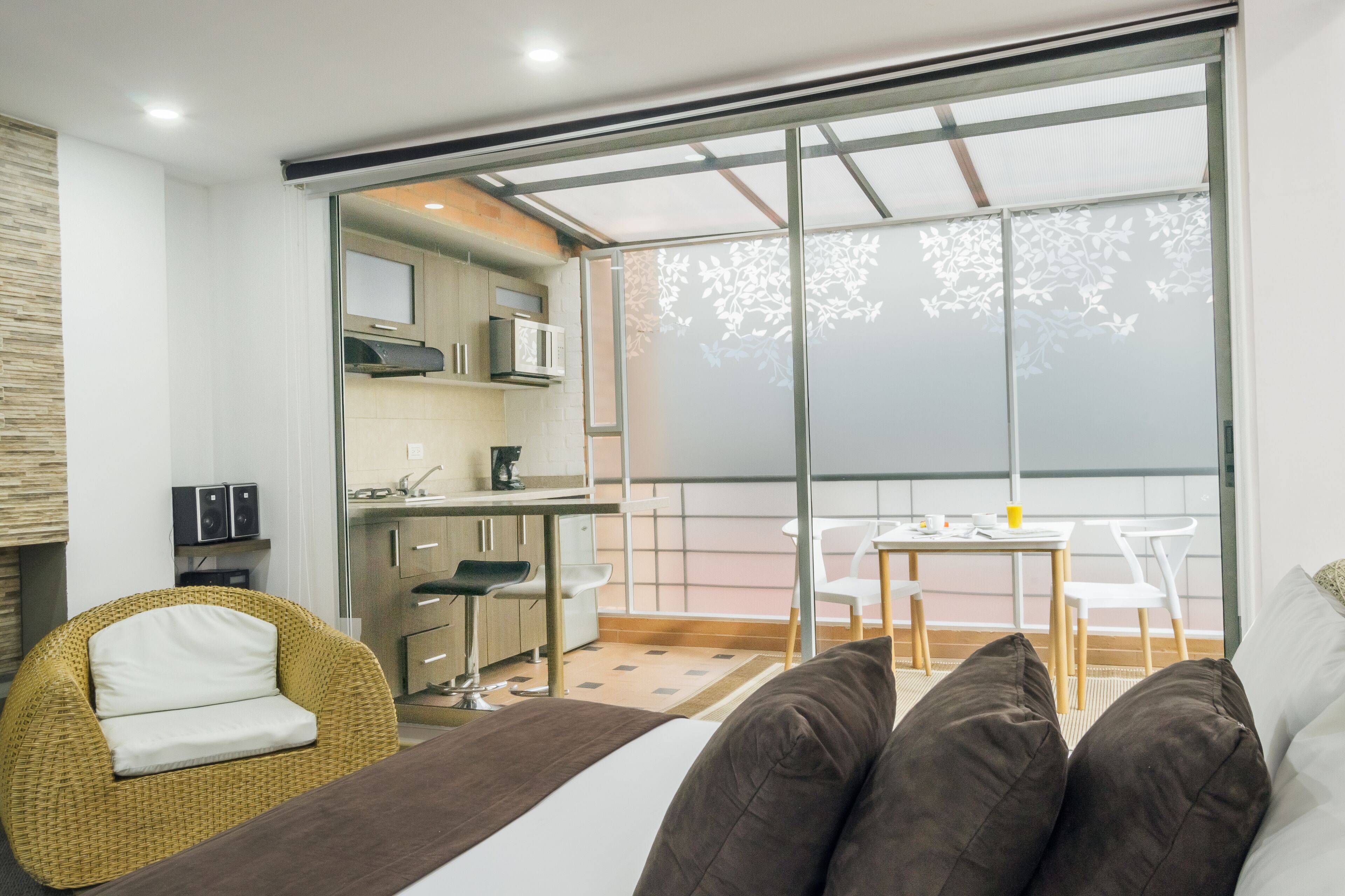 Una habitación moderna con cama, una silla de mimbre, un comedor con mesa y sillas, y una cocina con armarios y electrodomésticos.
