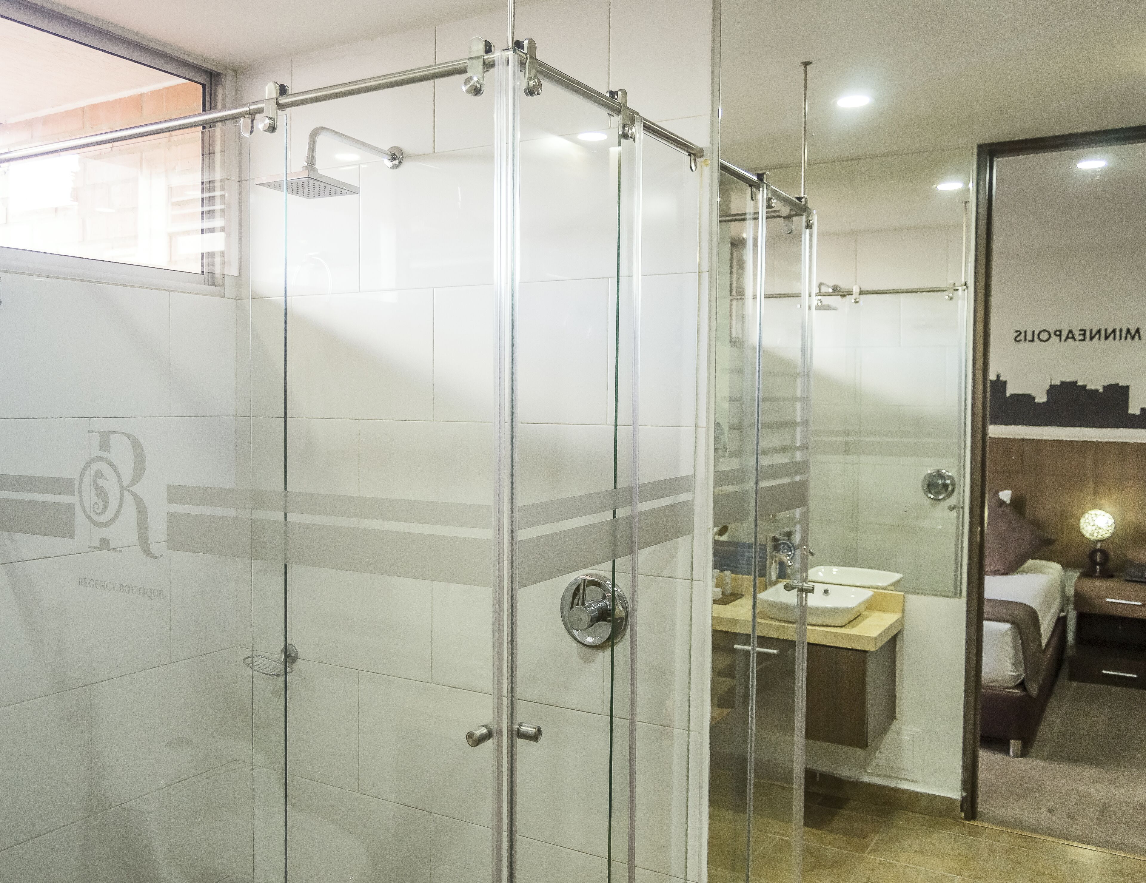 A modern bathroom with a glass-enclosed shower, a visible sink, and a bed in the adjacent room.