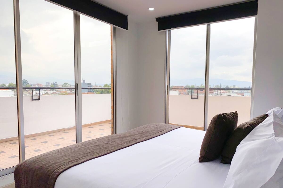 A hotel room with a large bed, two pillows, and a view of a cityscape through floor-to-ceiling windows.