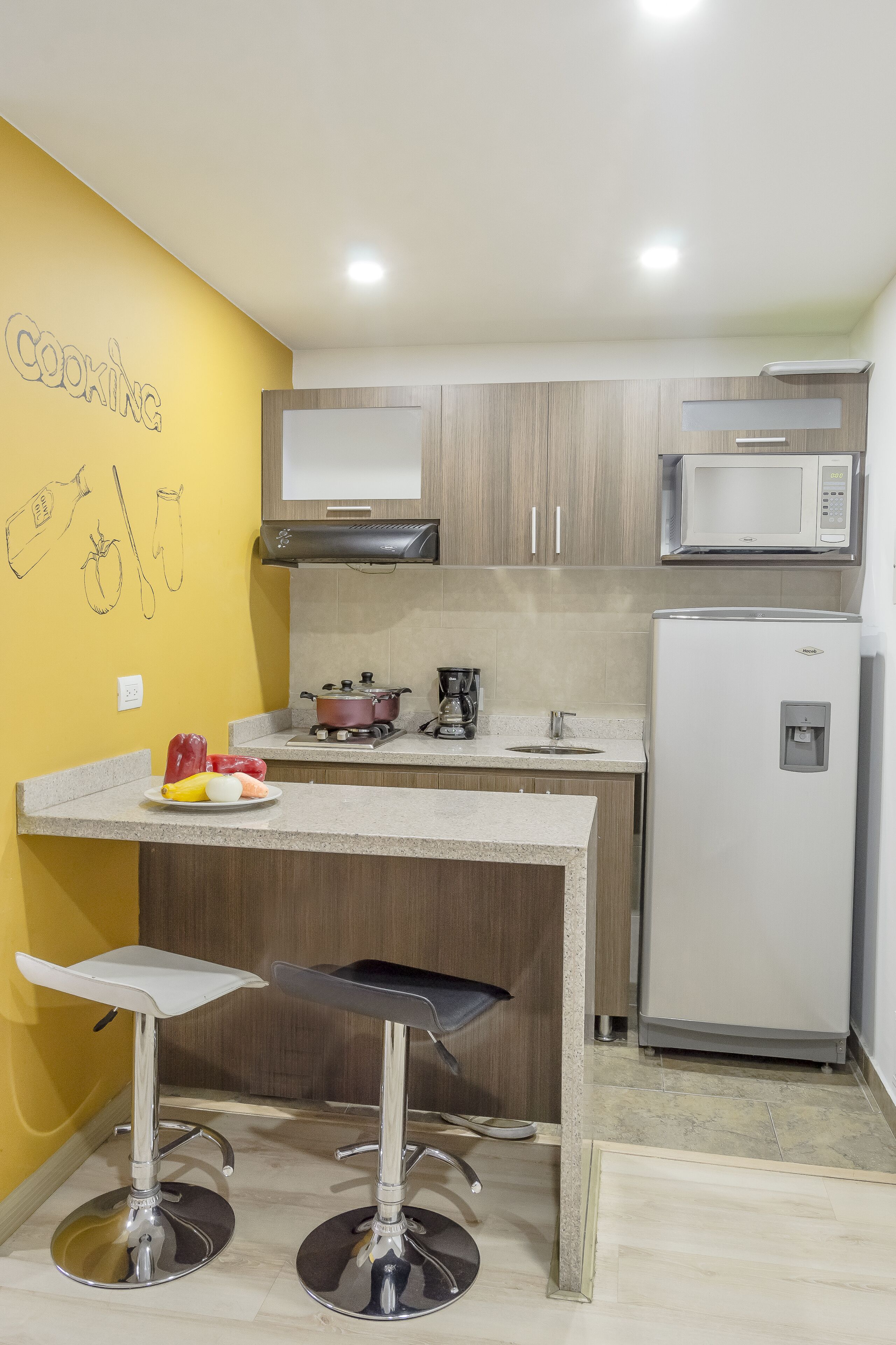 A kitchen with a yellow accent wall, a microwave, a refrigerator, a stove, and two bar stools.