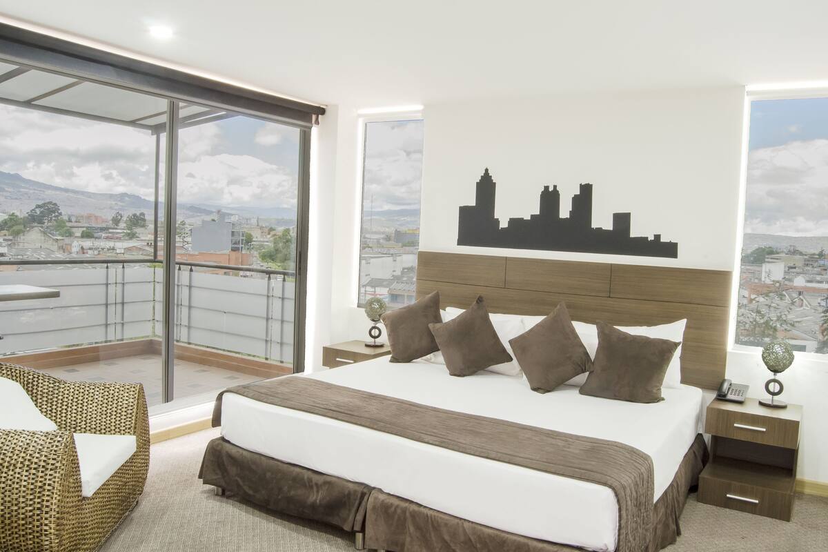 A modern hotel room with a large bed, a city skyline mural, a balcony with a glass door, and a view of the cityscape.
