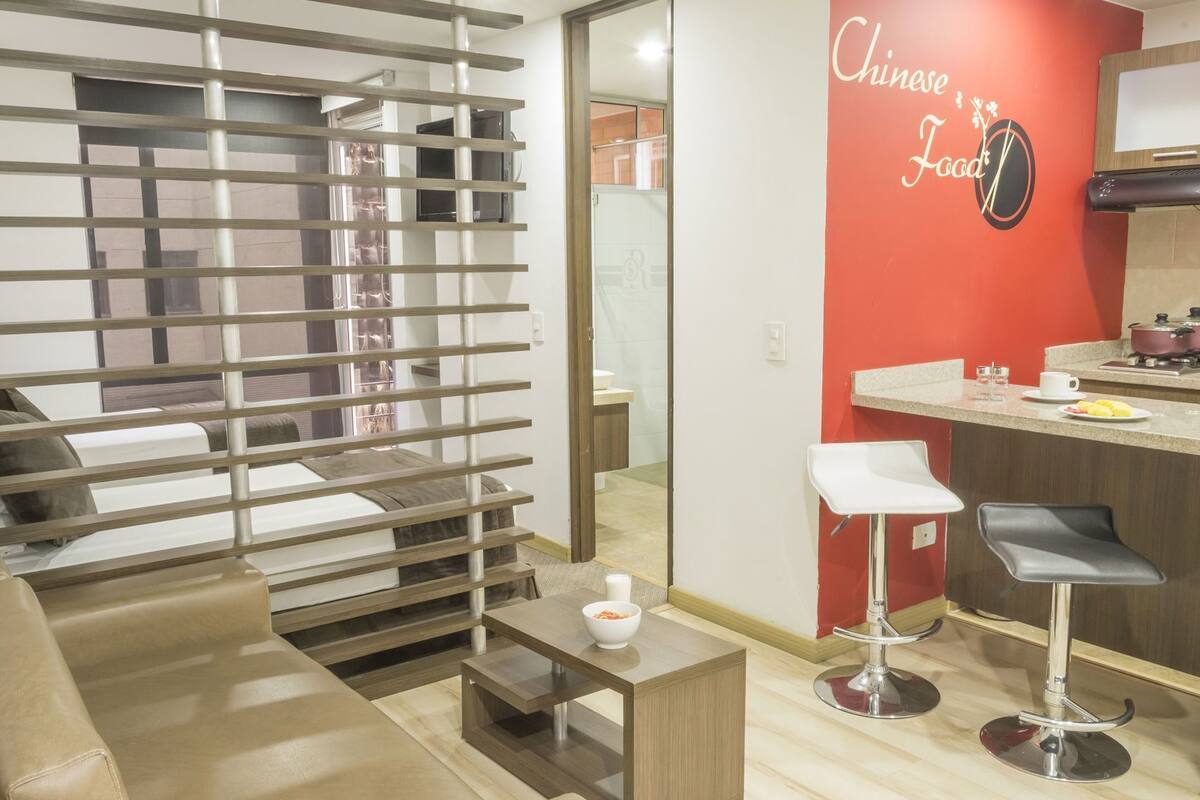 A modern living room with a sofa, a coffee table, a kitchen area with a red accent wall, and a dining area with two bar stools.