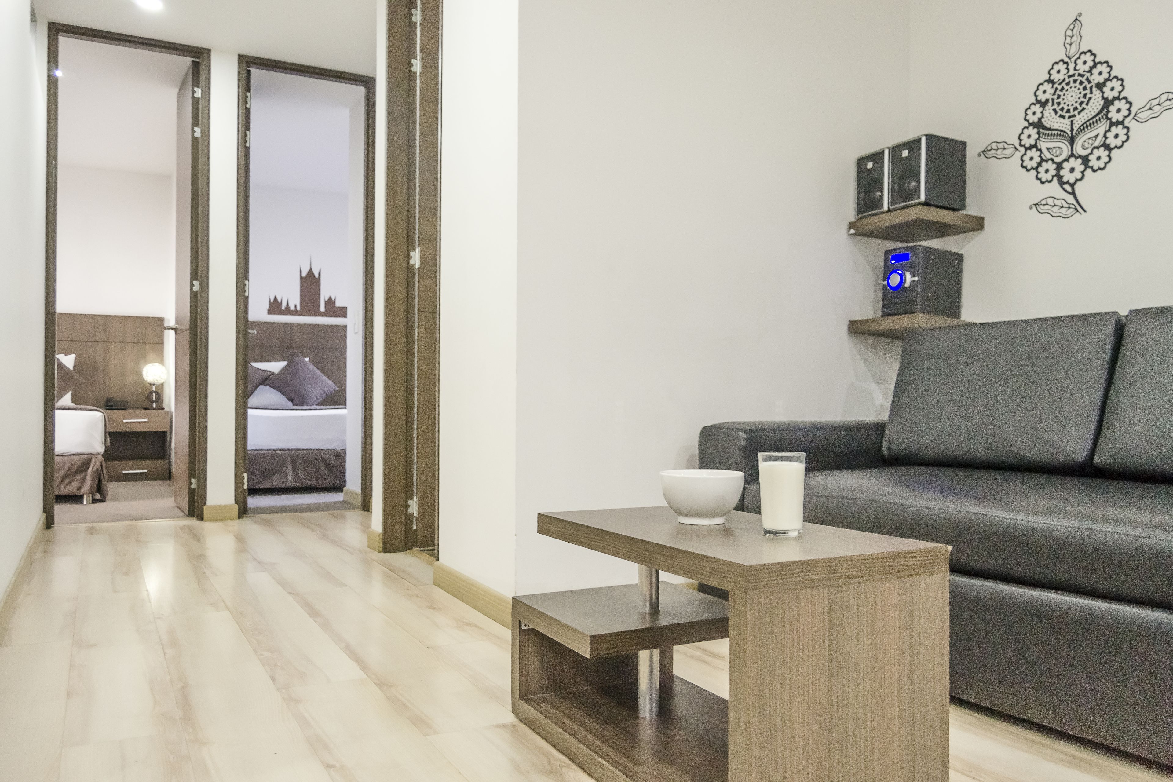 Un salon moderne avec un canapé gris, une table basse en bois et une petite table sur laquelle se trouvent un bol et un verre de lait. Deux haut-parleurs sont posés sur une étagère au-dessus du canapé.