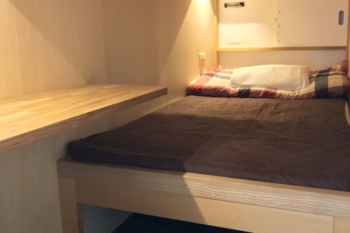 A small, single-bed room with a wooden headboard, a bedside table, and a wall-mounted shelf.