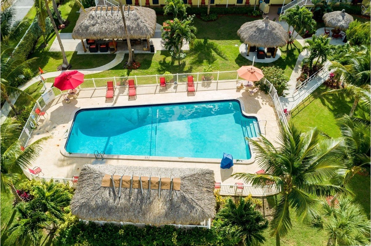 An aerial view of a resort with a central swimming pool, surrounded by lounge chairs, thatched-roof structures, and palm trees.