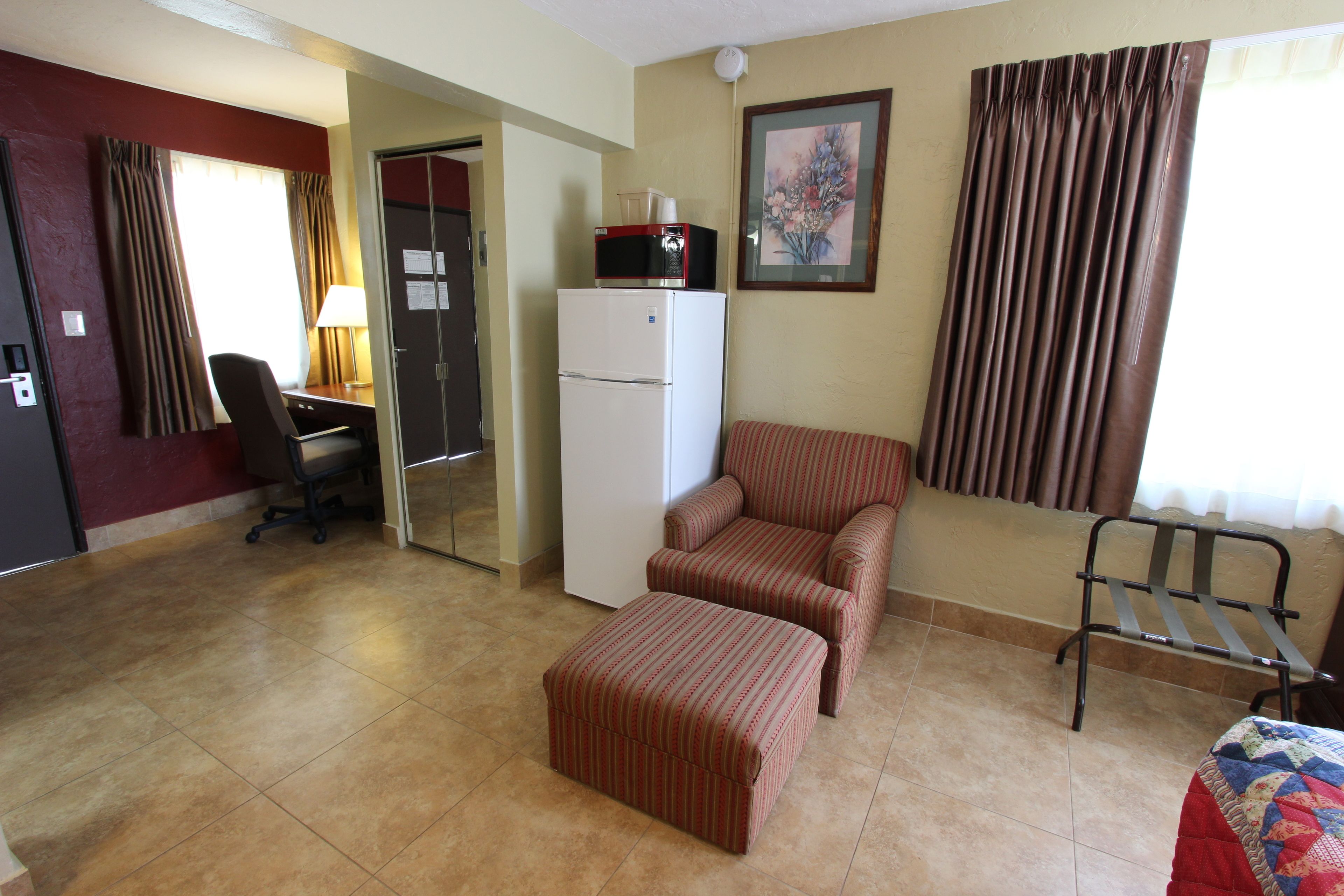A hotel room with a kitchenette, a desk, a chair, an ottoman, and a bed.