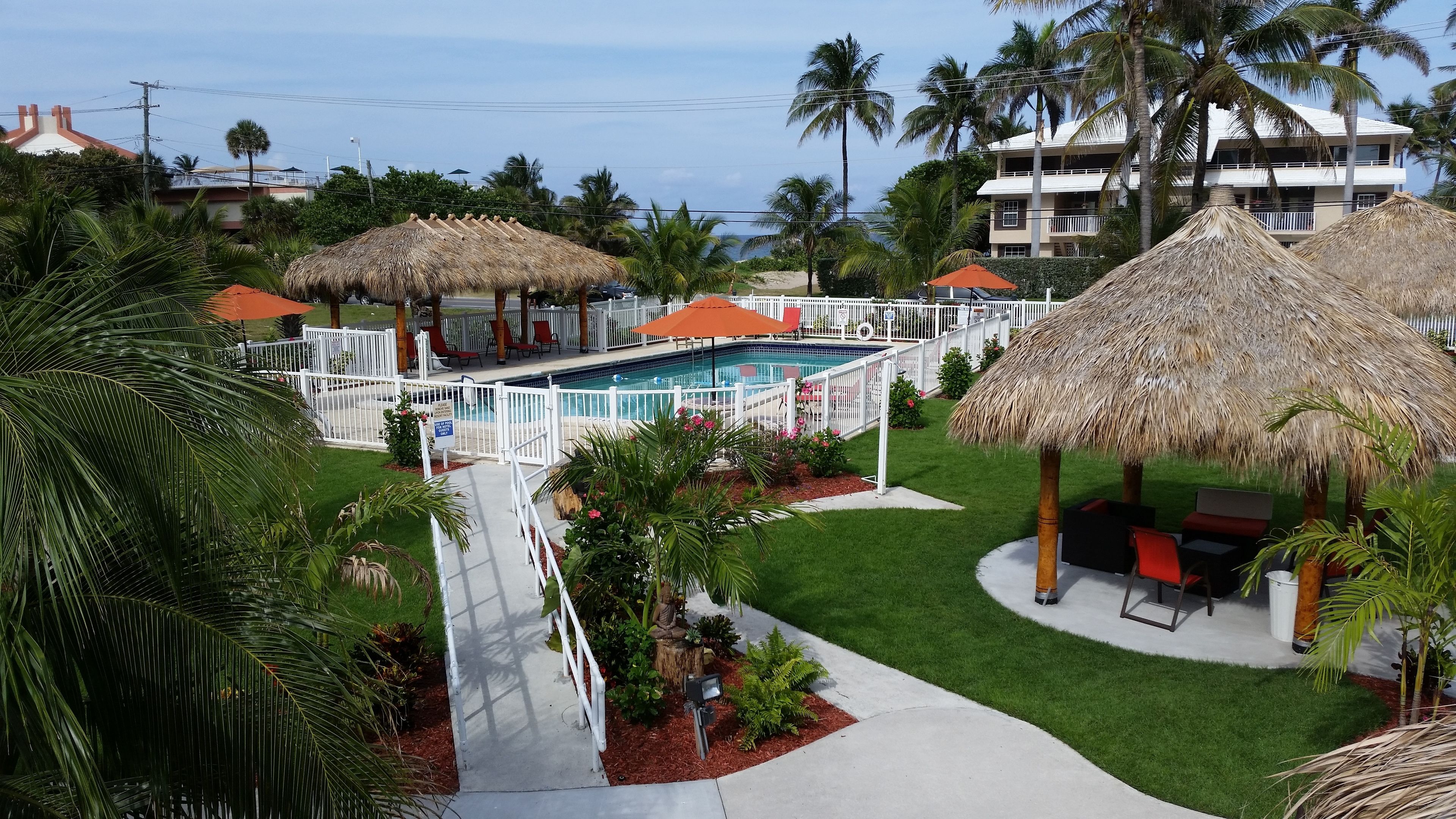 Zona de piscina con valla blanca, palmeras y sombrillas de paja.