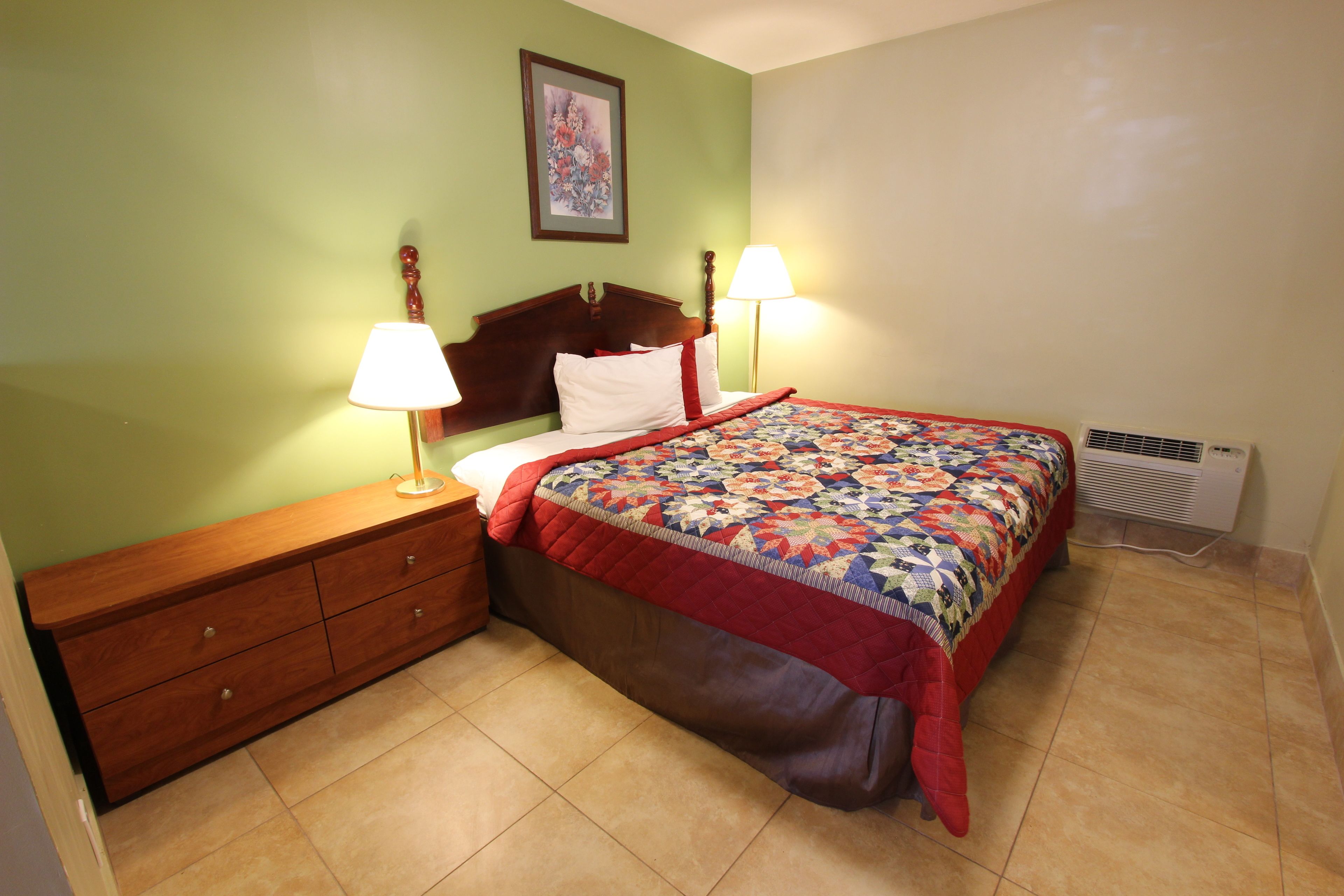 A bedroom with a wooden dresser, a bed with a quilt, two bedside lamps, and a wall-mounted picture.