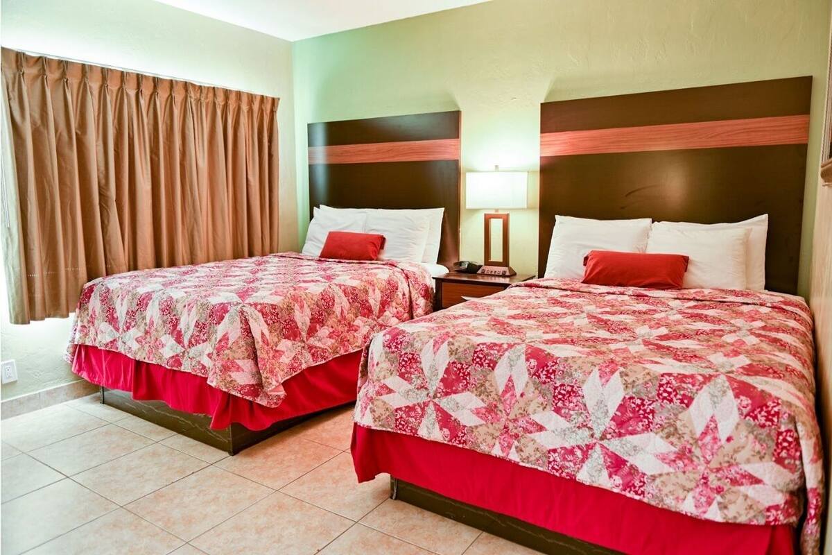 A hotel room with two beds, each with a floral-patterned bedspread and red accents. The room has a green wall and a brown curtain.