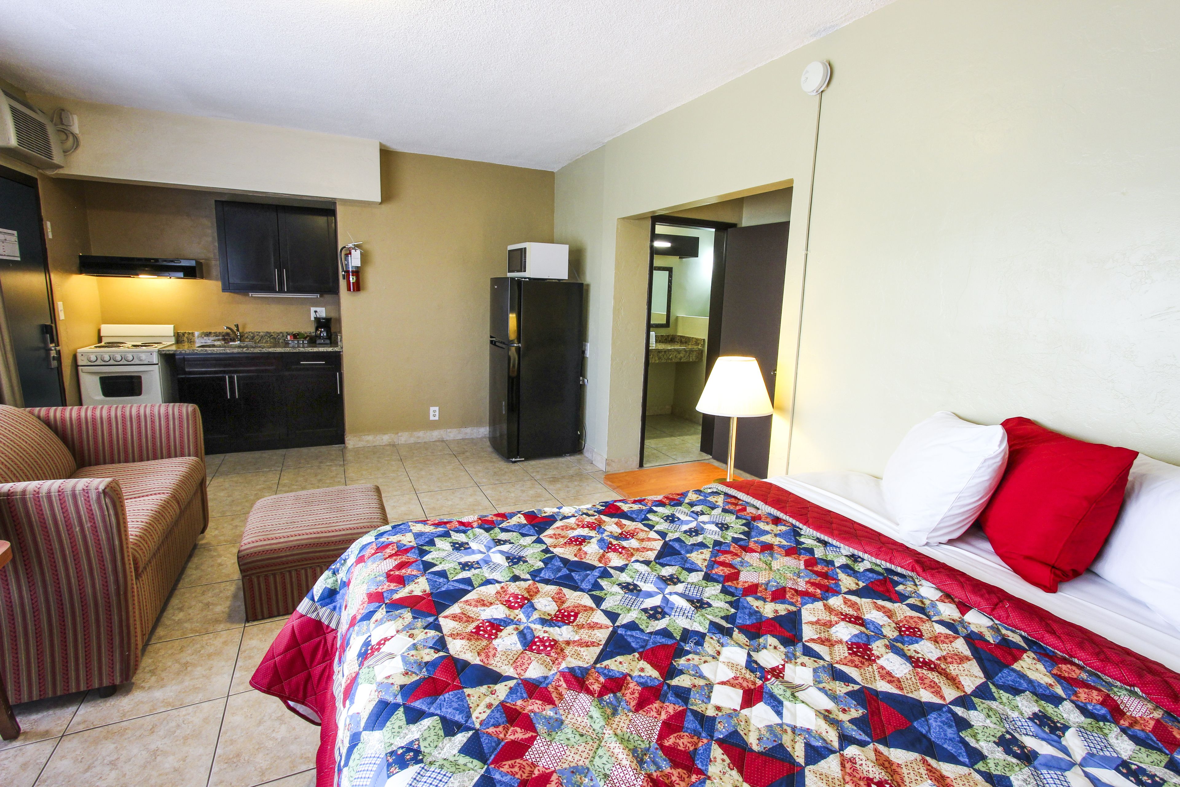 Una habitación de hotel con cama, kitchenette, un sillón a rayas y almohada roja.