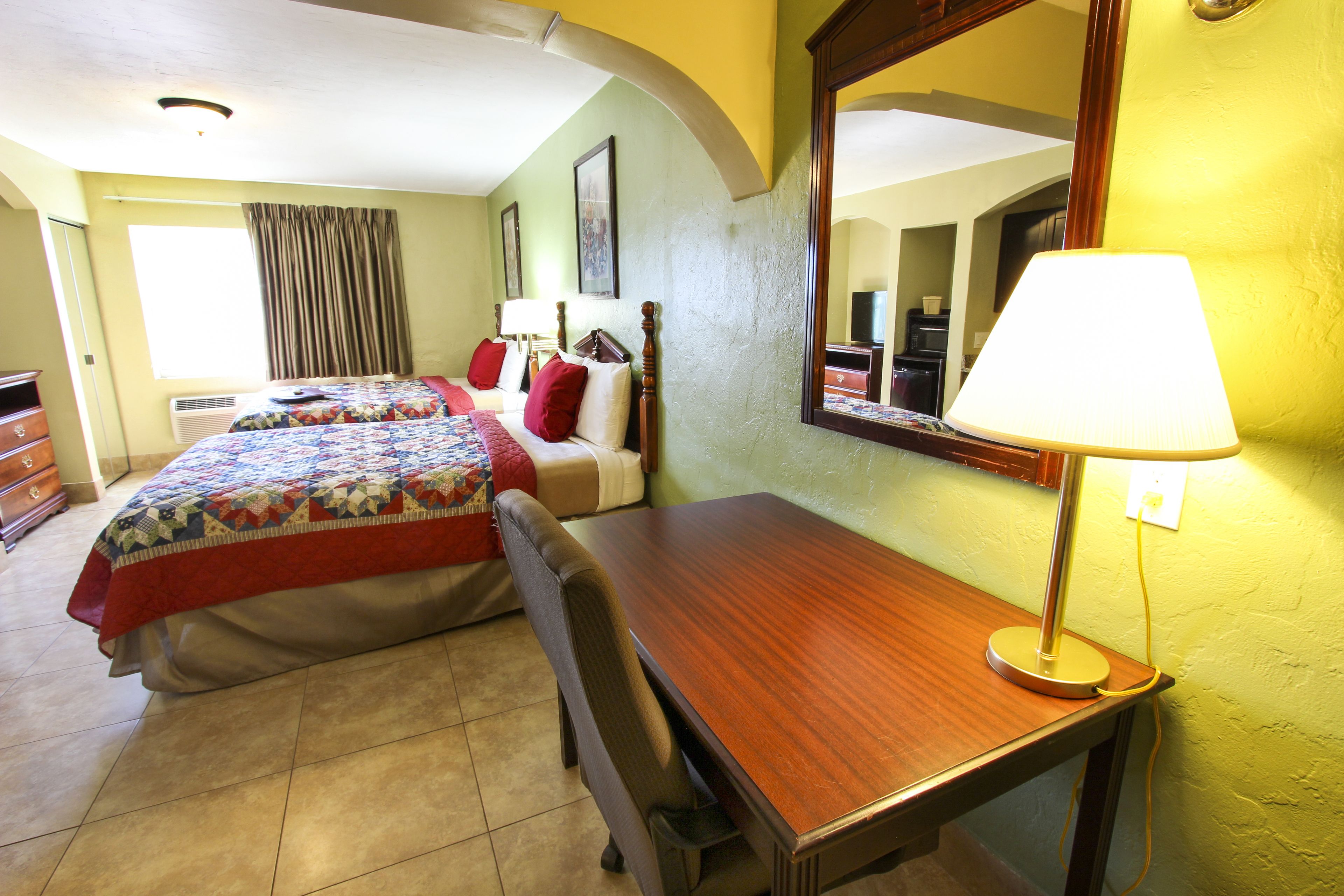 Une chambre d’hôtel avec deux lits, un bureau, une chaise, une lampe, un miroir et une fenêtre avec des rideaux.