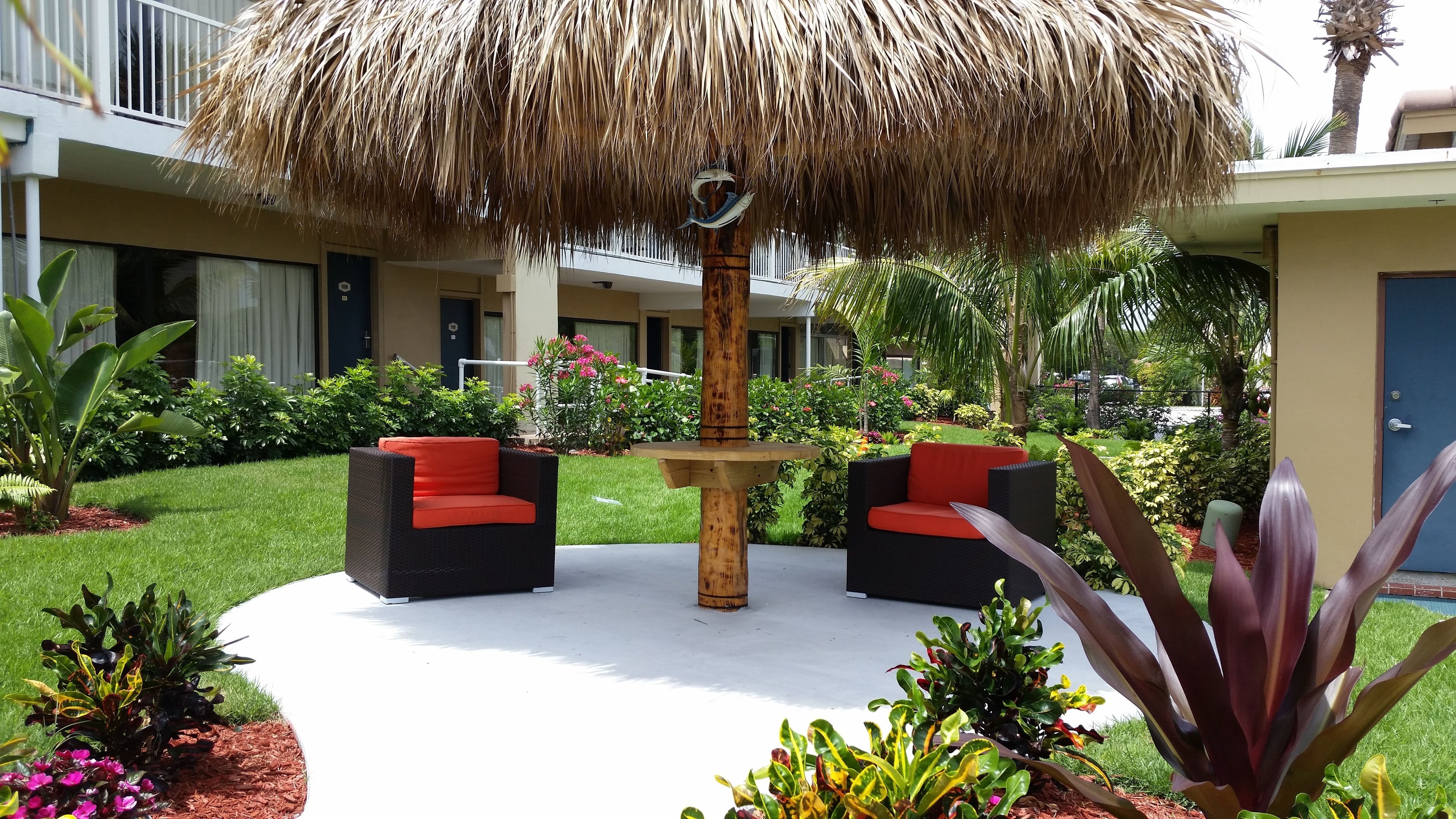 Un patio con un paraguas de paja, dos sillas de mimbre con cojines rojos y una variedad de plantas tropicales.