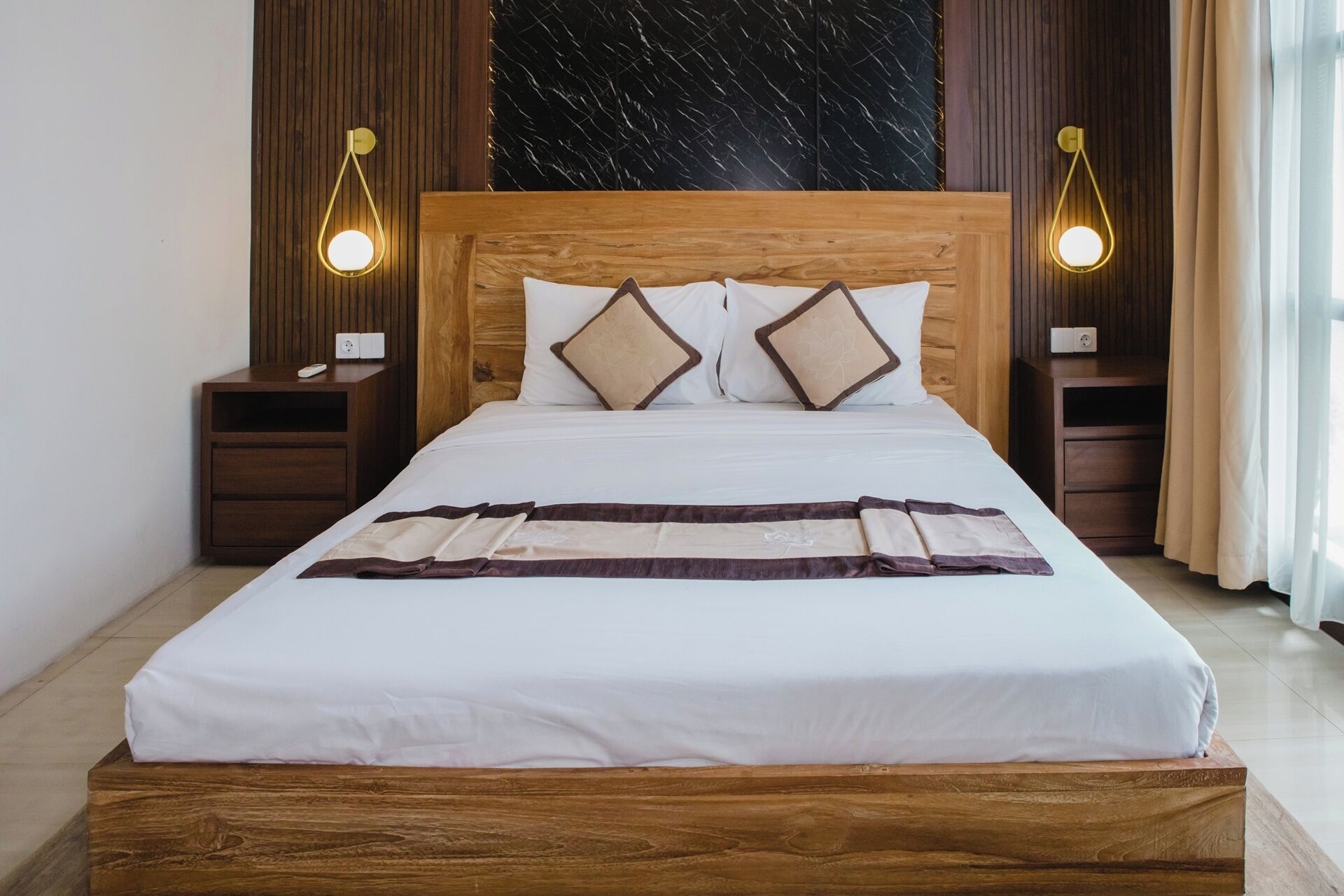 A neatly made bed with a wooden headboard, two bedside tables with lamps, and a marble-patterned wall behind.