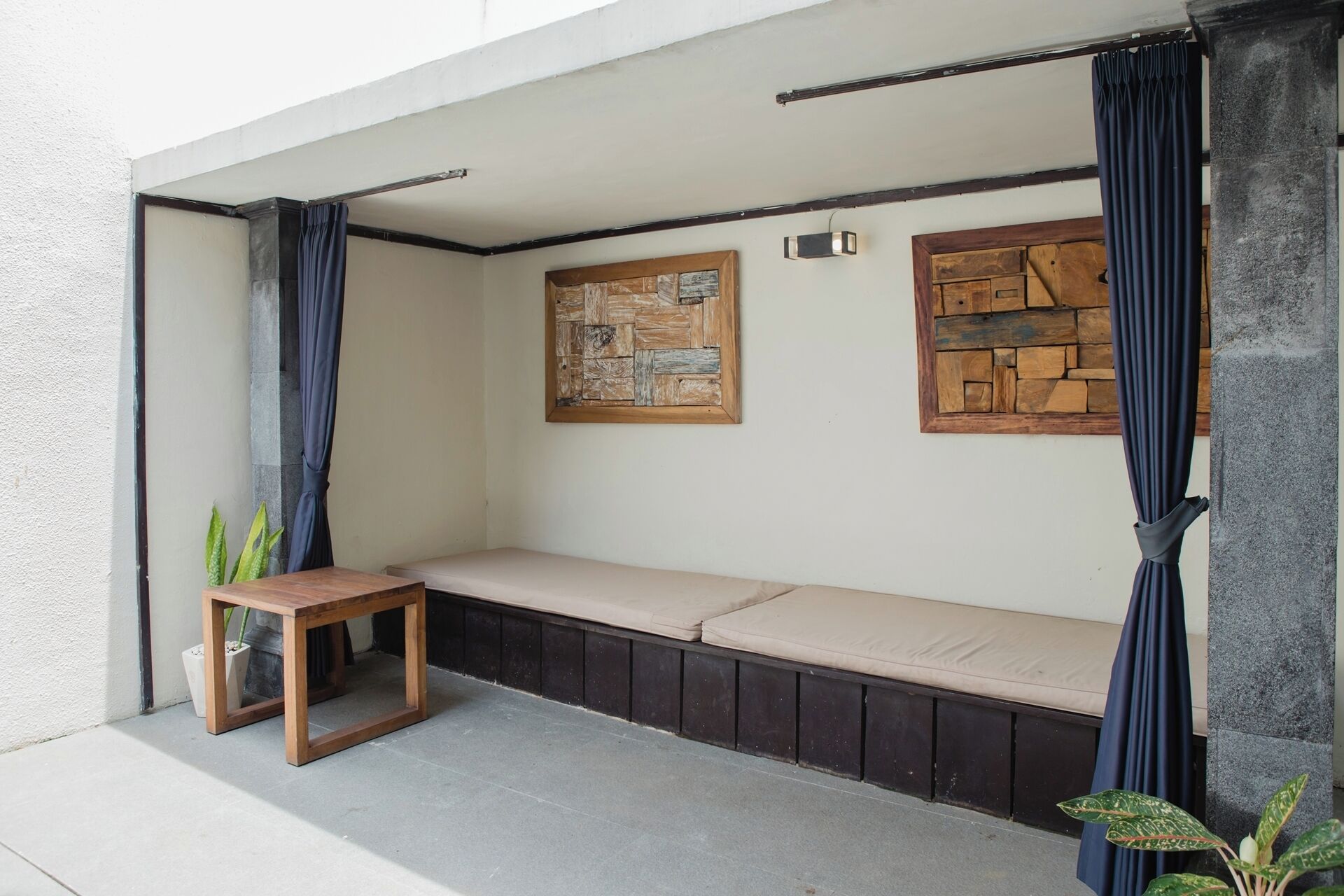 A small outdoor seating area with a bench and a table, surrounded by curtains and two wooden art pieces on the wall.