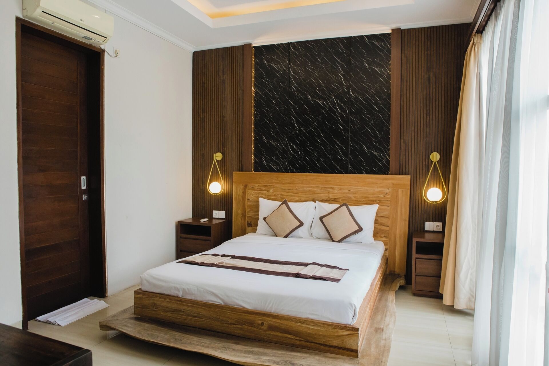 A modern bedroom with a wooden bed, two bedside tables, a marble-patterned wall, and a large window with curtains.