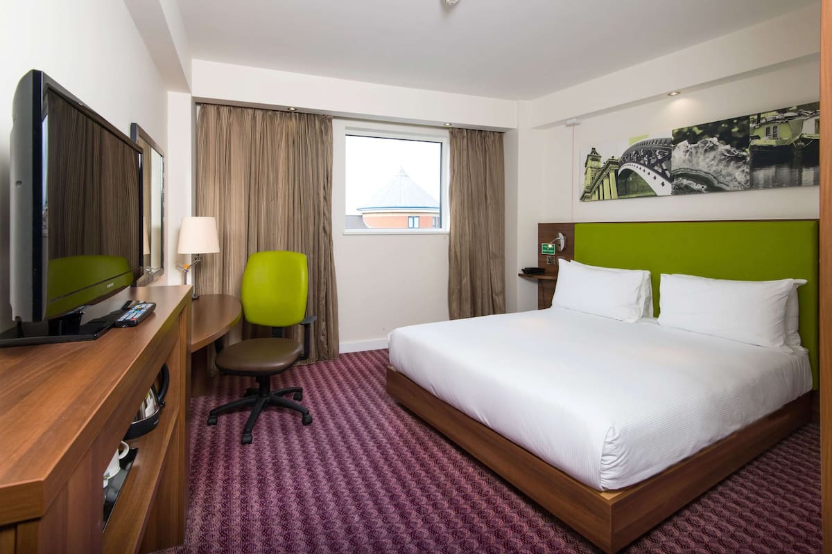 Habitación de hotel con una cama, un escritorio con computadora, una silla, una ventana con cortinas y un cuadro en la pared.