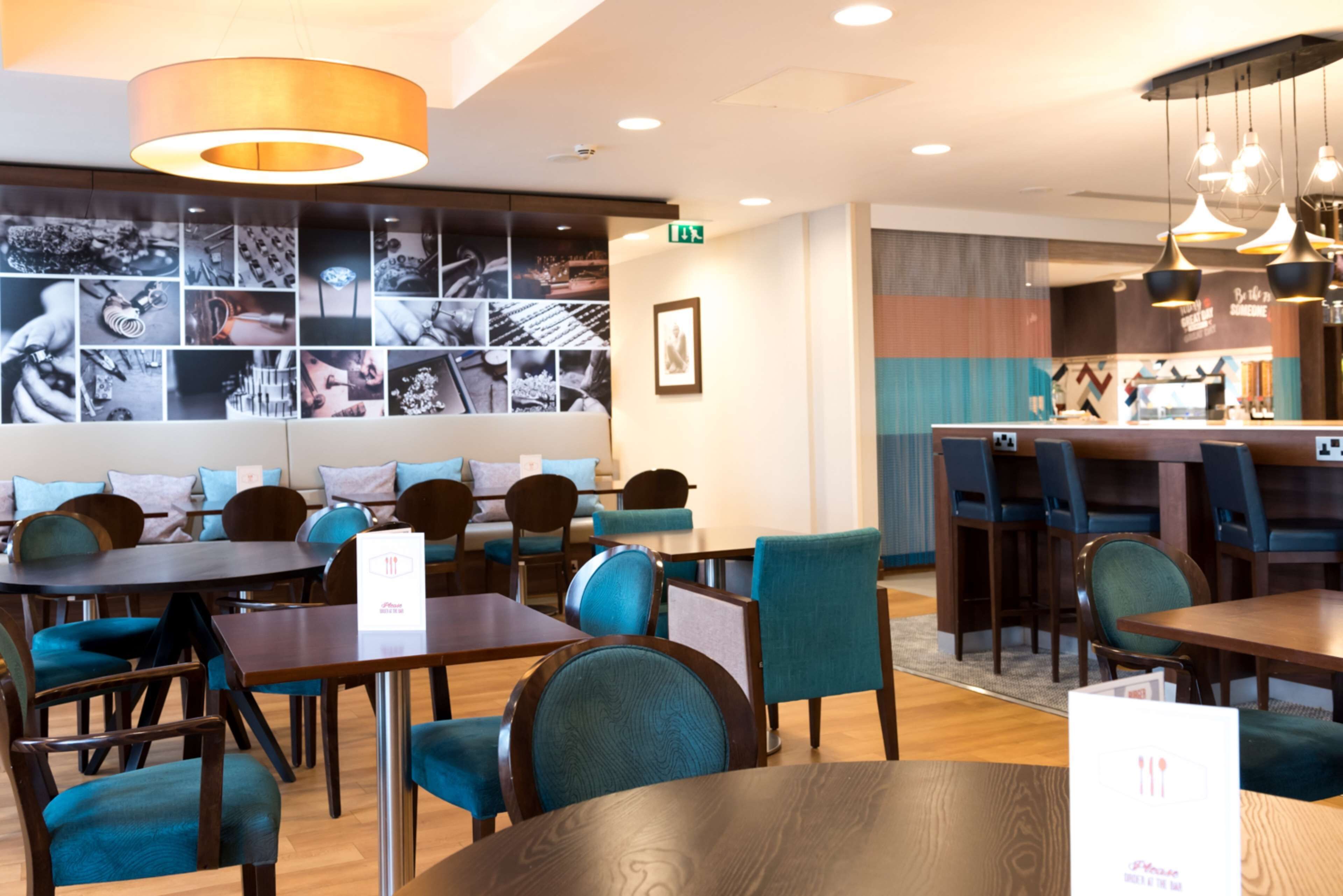 Un restaurante moderno con mesas de madera y sillas color turquesa, una pared decorada con fotografías en blanco y negro, y una zona de bar con luces colgantes.