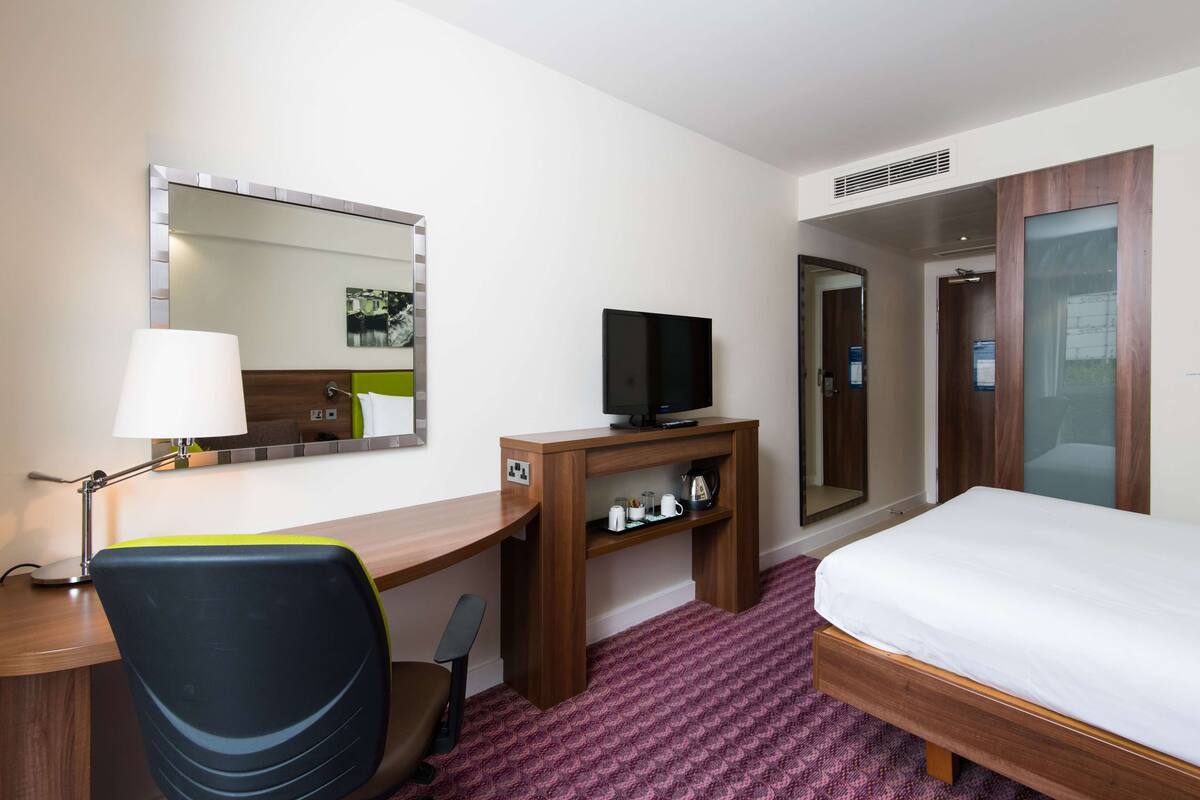 Habitación de hotel con cama, escritorio, silla, televisor y espejo.