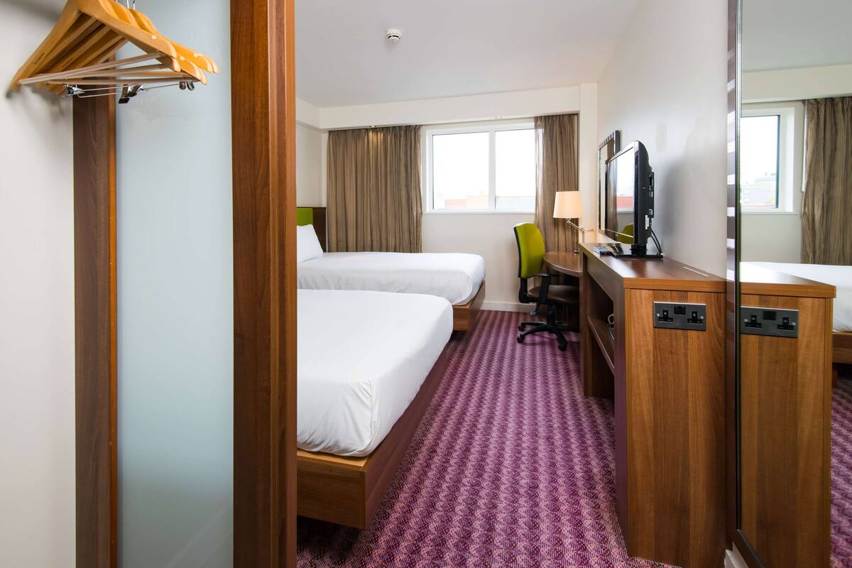 Habitación de hotel con dos camas individuales, un escritorio con silla, televisión y espejo.