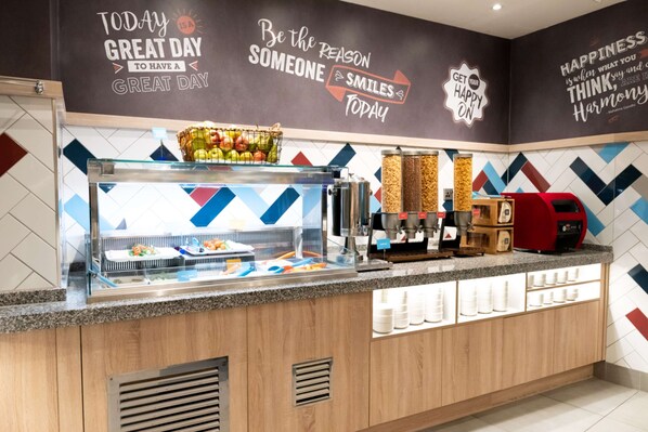 A breakfast bar with a fruit basket, coffee machine, and a wall with motivational quotes. - Hampton by Hilton Birmingham Jewellery Quarter (Birmingham)