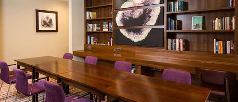 A conference room with a long wooden table, purple chairs, a bookshelf with books, and a framed picture on the wall.