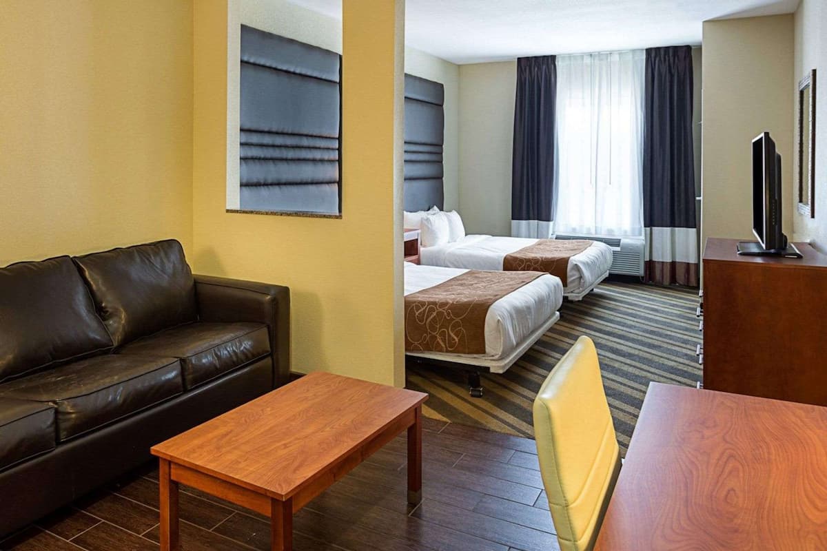 A hotel room with a leather sofa, a wooden coffee table, two beds with headboards, a television on a wooden stand, and a window with curtains.
