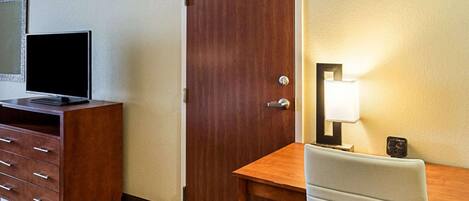 A hotel room with a wooden desk, a chair, a television on a cabinet, a door, and a wall-mounted lamp.