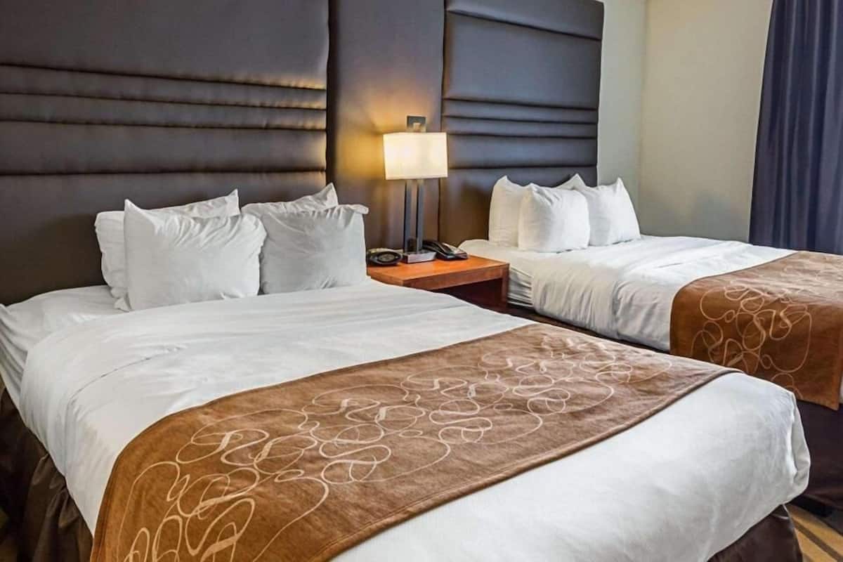 A hotel room with two beds, a headboard, a bedside table with a lamp, and a curtain.