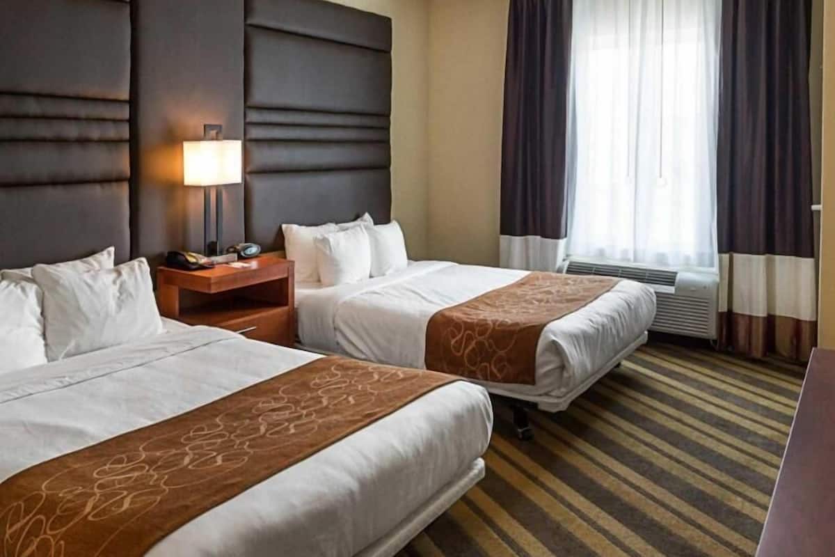 A hotel room with two beds, a nightstand with a lamp, an air conditioning unit, and a window with curtains.