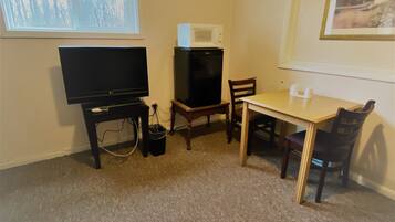 A small room with a television, a microwave on a stand, a table with chairs, and a window with a view of trees.