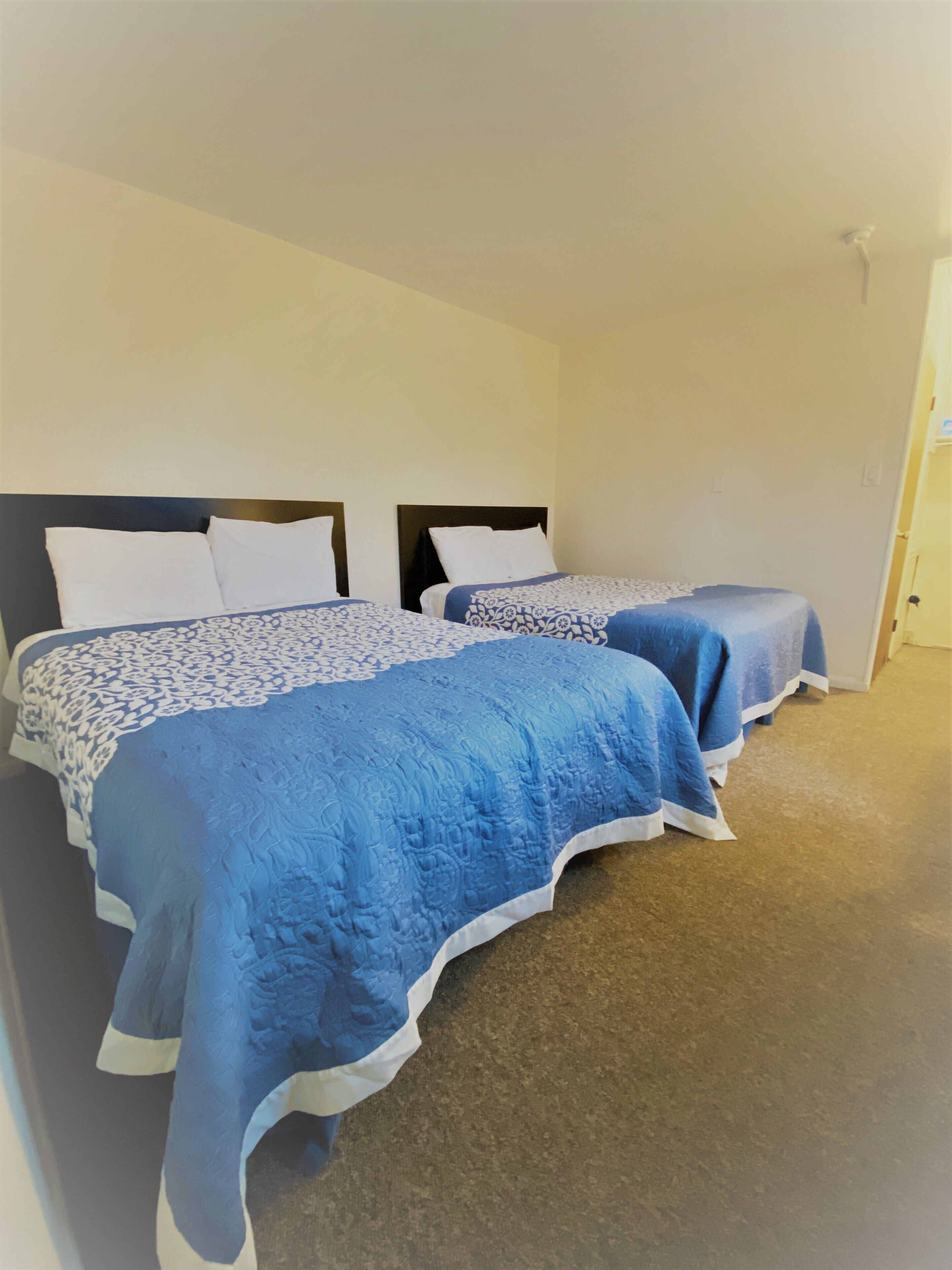 Dos camas individuales con cabeceras, ropa blanca de cama y mantas azules en una habitación con paredes color beige y suelo de alfombra.