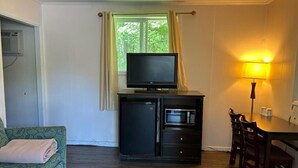 Une pièce de taille modeste, équipée d’un téléviseur posé sur un meuble, d’un four à micro-ondes, d’un petit réfrigérateur, d’une table à manger avec des chaises et d’un canapé avec une couverture.
