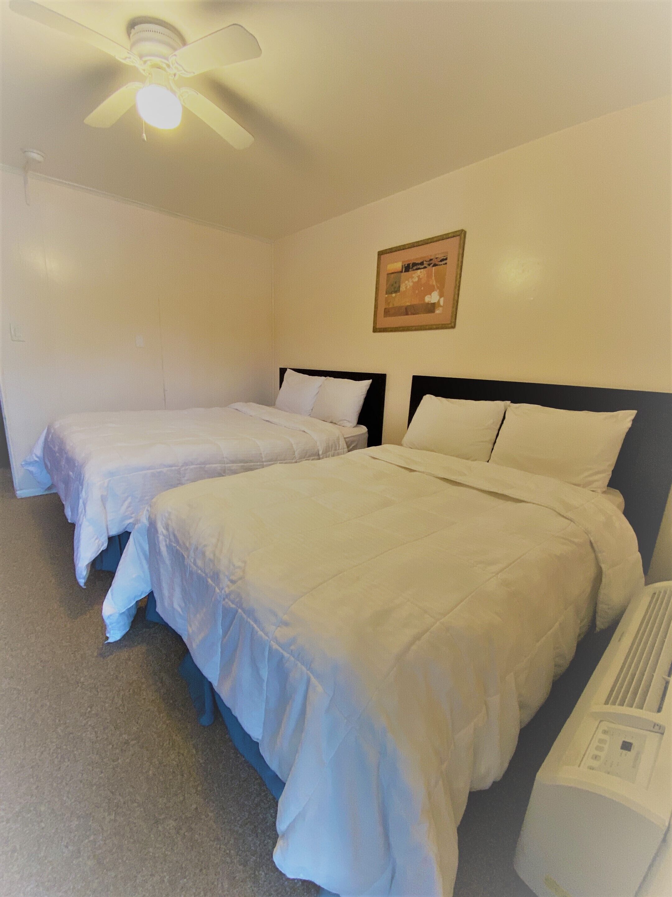 A hotel room with two single beds, a ceiling fan, a wall-mounted air conditioner, and a framed picture.