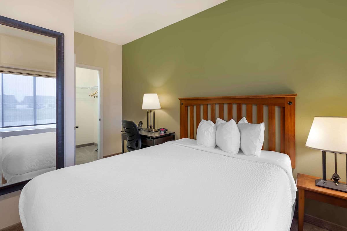A hotel room with a large bed, a desk with a chair, a nightstand with a lamp, and a mirror.