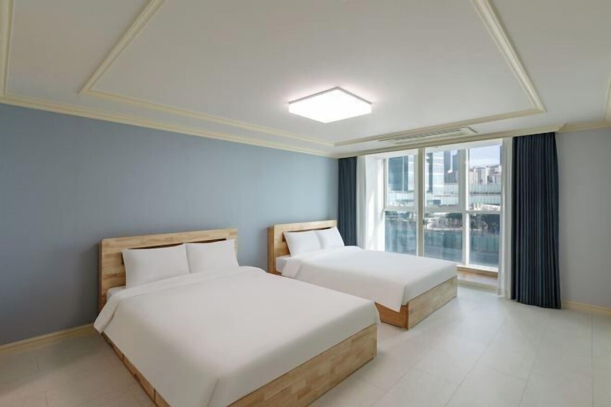 A hotel room with two beds, a large window with a city view, and a modern ceiling light.