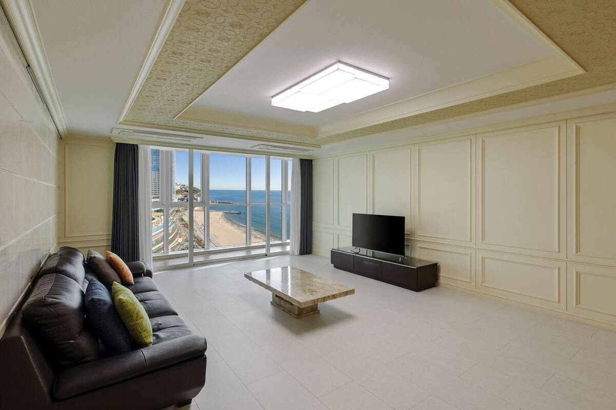 Un salón amplio con un gran ventanal que ofrece vistas a la playa, un sofá de cuero con cojines, una mesa de centro y un televisor de pantalla plana sobre un mueble.